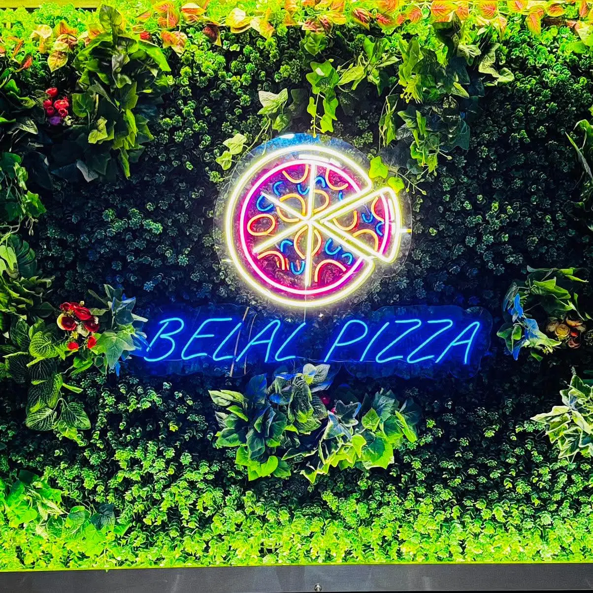 The blue neon Belal Pizza sign on a wall of green foliage